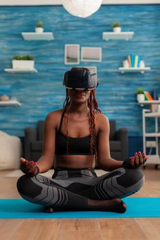 Black woman experiencing virtual reality training body and mind meditating in lotus pose sitting on yoga mat in home living room for calm, healthy harmony lifestyle dressed in sportwear.