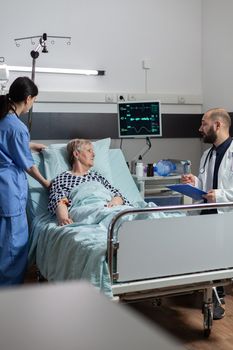 Doctor talking with elderly patient sitting next to bed in hospital room, giving expertise for treatment. Senior woman breathing with help from oxygen mask laying in bed.