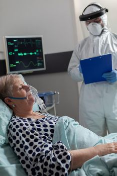 Sick senior woman inhale and exhale through oxygen mask laying in hospital bed during global pandemic with coronavirus. Doctor dressed in coverall pee suit to prevent infection with covid-19.