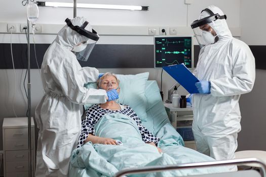 Medical team in clinic wearing coverall ppe and face shield during patient visit in hospital room in time of coronavirus out break to prevent infection. Uconscious senior woman breathing with help from face mask.