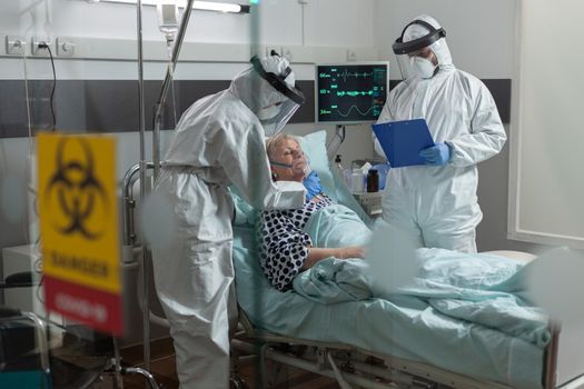 Medical nurse dressed in ppe suit putting oxygen mask to senior patient, in the course of global pandemic with coronavirus and doctor is taking notes on clipboard.