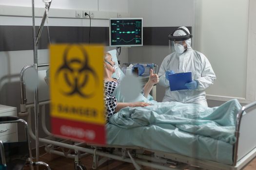 Doctor wearing ppe protection suit against contamination with covid-19, in hospital room during visit at senior woman laying in bed breathing with help from oxygen mask. and getting intravenous medicine.