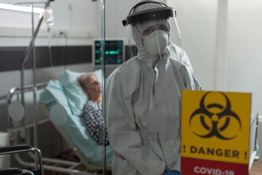 Portrait of medical nurse dressed in pee suit in hospital room looking at camera, with senior woman patient laying in bed breathing heavely with help from oxygen mask.