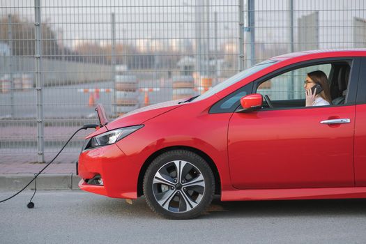 Woman using smart phone and waiting power supply connect to electric vehicles for charging the battery in car. Positive young girl talking on the phone sits in electric car and charging