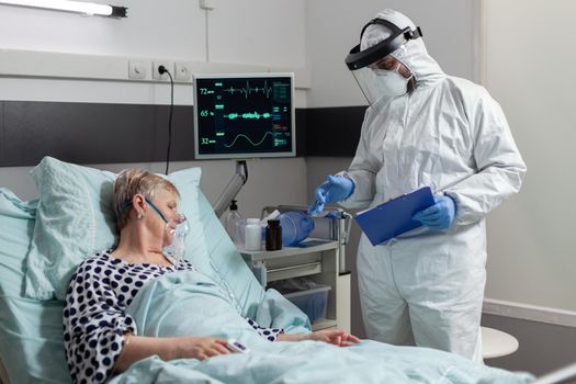 Sick senior patient getting intravenous medicine from iv drip bag laying in bed, inhale and exhale through oxygen mask, during coronavirus pandemic. Doctor wearing ppe suit.