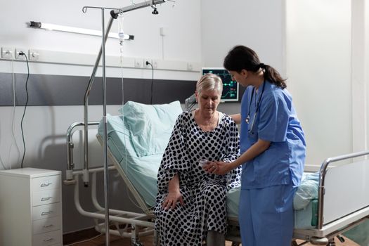 Medical nurse in scrubs helping senior woman in hospital room giving support to get up for treatment. Oxymeter attached on patient showing oxygen saturation and blood pressure. Getting intrevenous medicine from iv bag.