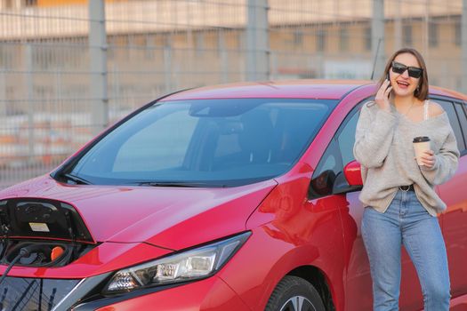 Stylish girl stands with phone near her red electric car and waits when vehicle will charged. Connecting the Charger Plug of an Electric Car