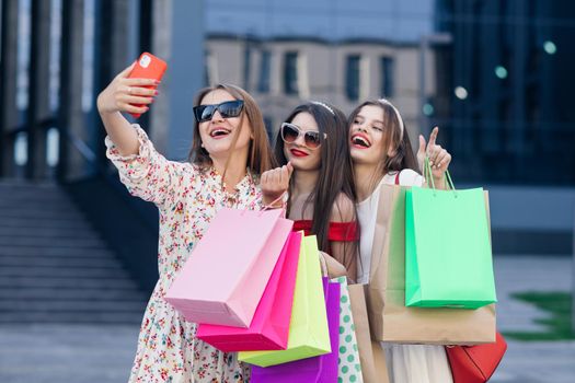 Group of young beautiful girls in casual clothes with sunglasses, makeup, hair hoop and colored shopping bags making selfie after successful shopping.