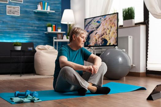Senior woman sitting in lotus position on yoga mat training body muscles slimming weight after watching online fitness lesson using laptop computer. Pensioner stretching in living room