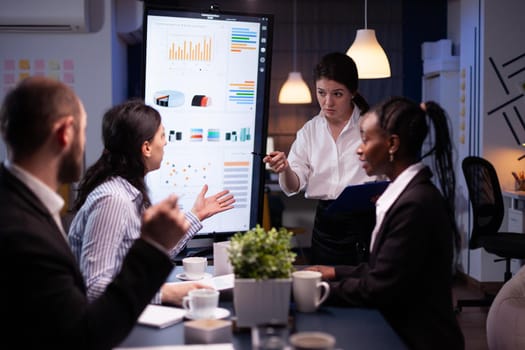 Multi-ethnic businesspeople discussing financial company solution sitting at conference table in meeting room. Diverse teamwork overworking at management strategy using tv late at night.