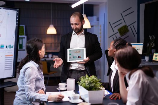 Overworked businessman showing financial graphs presentation using tablet brainstorming company ideas. Workaholic diverse multi-ethnic businesspeople overworking late at night in office meeting room