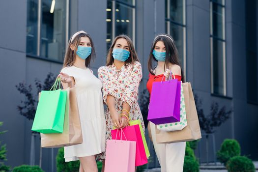 Group of young beautiful women in casual dresses, top and pants wearing masks to protect coronavirus pandemic standing in front of the shopping mall with colored bags in hands.