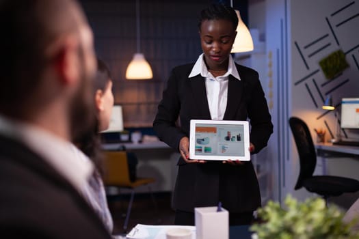 Focused african american businesswoman showing company graphs on tablet working in office meeting room late at night. Diverse multi-ethnic teamwork brainstorming corporate strategy ideas