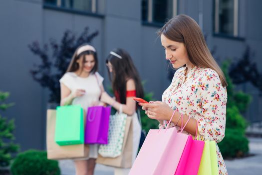 sale, consumerism, technology and people concept - happy young women with smartphones and shopping bags