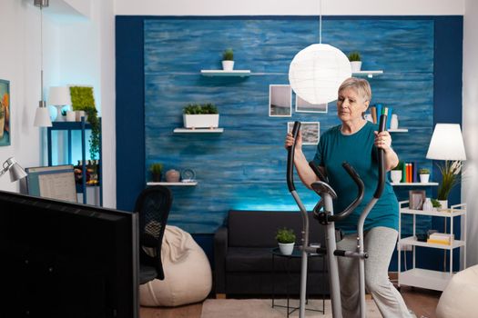 Focused senior woman working at legs muscle resistance cycling bicycle machine in living room during aerobics healthy workout. Pensioner watching fitness video on televison for well being