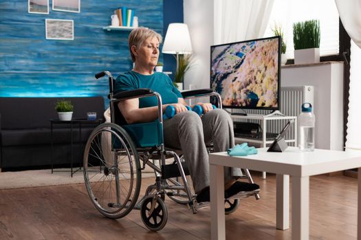 Disability senior woman in wheelchair looking at gymnastic online video on tablet computer exercising body muscle using dumbbells. Invalid pensioner wearing sportswear working arms resistance