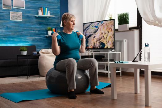 Focused senior man exercising body resistance workout doing arms exercise using dumbbell sitting on fitness swiss ball in living room. Pensioner watching online wellness training on laptop