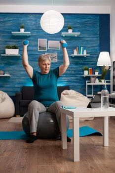 Senior woman in retirement raising hands doing arms exercise using dumbbells sitting fitball training body resistance. Pensioner workout pilates exercises watching online fitness video on tablet