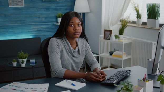 Portrait of cheerful black woman looking at camera while sitting at desk table in living room during homeschooling. Student smiling, study browsing high school news on computer