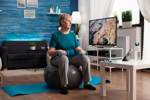 Focused senior woman in sportswear watching online fitness workout on tablet sitting on swiss ball exercising body muscle. Pensioner training arms resistance using aerobics dumbbells in living room
