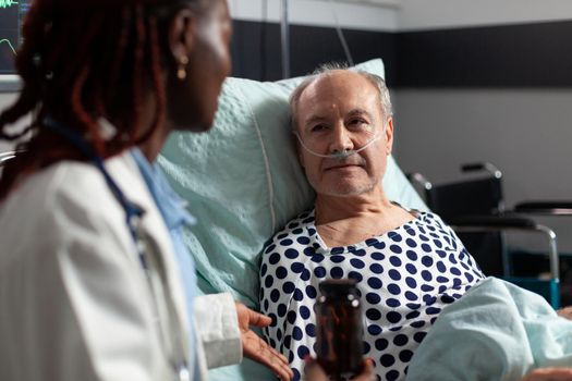 Close up of sick unwell senior man patient laying in hospital bed. listening doctor specialist discussing about treatment holding pills bottle reassuring full recovery. Man breathing with help from oxygen tube.