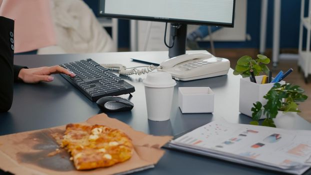 Closeup of businesswoman sitting at desk in front of computer eating pizza slice while talking at landline with remote company manager. Takeout order food delivery in corporate job place