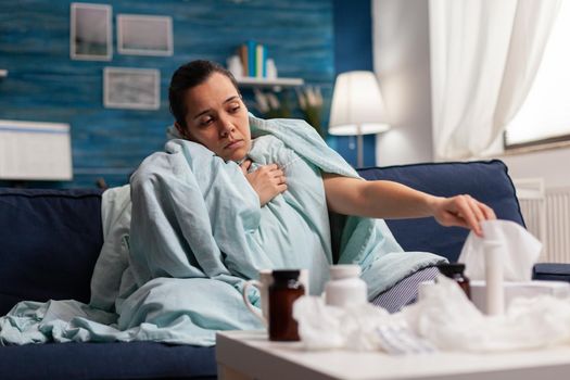 Sick woman wrapped in blanket at home with virus infection illness fever sickness flu cold. Caucasian young adult suffering from temperature fever headache on couch using tissues