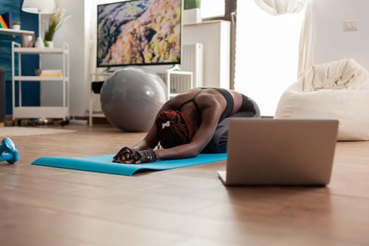 Black woman sitting on yoga mate doing stretching relaxing body after watching online training looking at laptop in home living room, on the floor for health wellbeing.