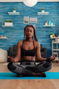 Black woman training body and mind meditating in lotus pose keeping eyes closed, sitting on yoga mat in home living room for calm, healthy harmony lifestyle dressed in sportwear.