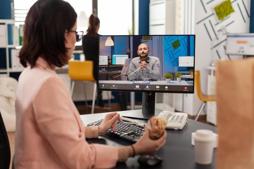 Entrepreneur woman sitting at desk in company office eating sandwich during online videocall conference meeting discussing financial strategy. Takeout order food delivery in corporate job place