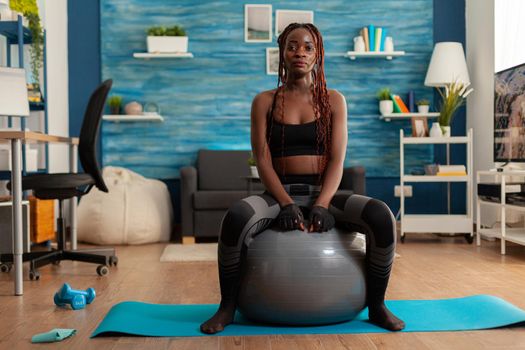 Sportive active strong black woman sitting on stability ball resting, after intense training in home living room. Slim fit african dressed in sportwear leggings.