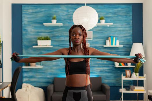 Black strong fit woman standing in living room working out shoulders using resistance band dressed in sportive sportwear. Active african exercising with rubber band.