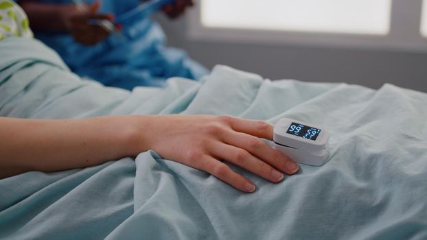 Close up of sick woman resting in bed in hospital ward with finger heart rate monitor puls while medical team monitoring heartbeat after blood test. Patient with oximeter waiting for illness treatment