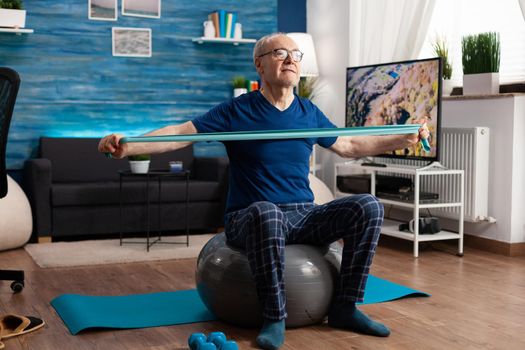 Cheerful retired senior man exercising arm muscles using resistance elastic band practicing aerobic exercise. Pensioner sitting on swiss ball in living room working at body healthcare