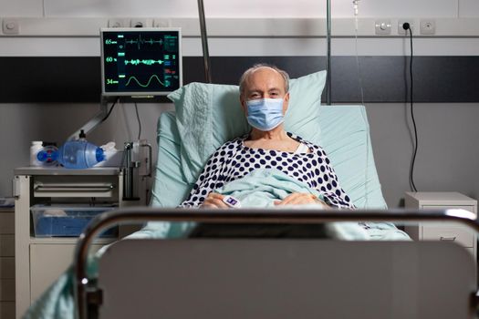 Portrait of sick senior man patient with chirurgical mask resting in hospital bed, with iv drip attached on hand having oximeter attached on finger. Modern medical equipment.