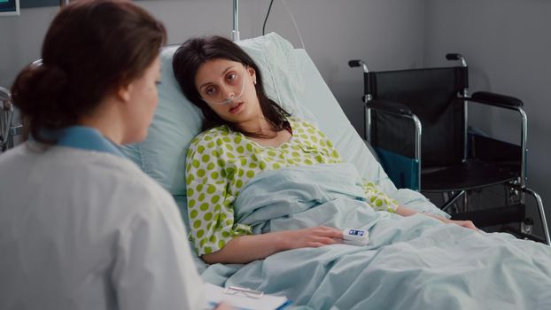 Doctor practitioner writing sickness symptom on clipboard checking pulse using medical oximeter during recovery appointment in hospital ward. Sick woman patient resting in bed recovery after surgery