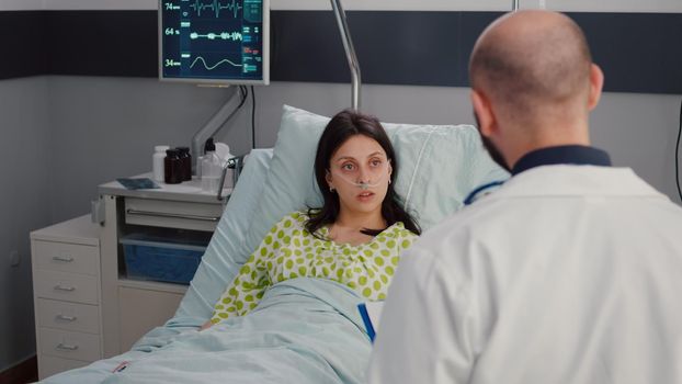 Closeup of sick woman with nasal oxygen tube resting in bed. Patient discussing with practitioner doctor about medical healdcare expertise checking disease test results working in hospital ward