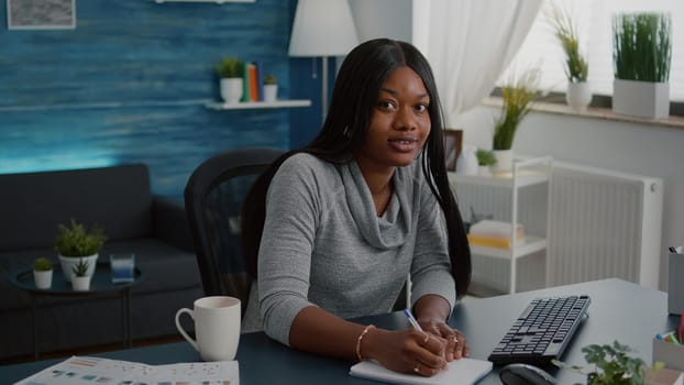 Student with black skin browsing online courses writing school homework on notebook during online webinar education. Young woman sitting at desk table in living room typing lifestyle information