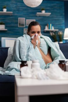 Sick woman wrapped in blanket at home with virus infection illness fever sickness flu cold. Caucasian young adult suffering from temperature fever headache on couch using tissues