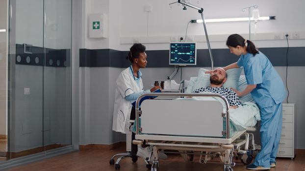 Afro american practitioner monitoring sick man analyzing sickness symptom during recovery appointment in hospital ward. Patient resting in bed while assistant arrange to sit comfortable