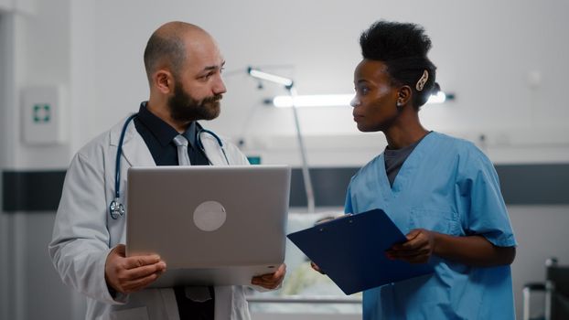 Assistant with black skin and practitioner doctor in medical uniform monitoring sickness symptom working in hospital ward. In background woman physician checking sick patient