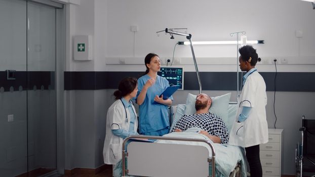 Doctors team checking sick man discussing sickness symptom monitoring medical recovery in hospital ward. Patient resting in bed waiting disease treatment during rehabilitation appointment