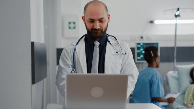 Front view of practitioner doctor typing illness expertise on computer while in background black nurse discussing healthcare treatment. Hospitalized patient having respiratory disorder