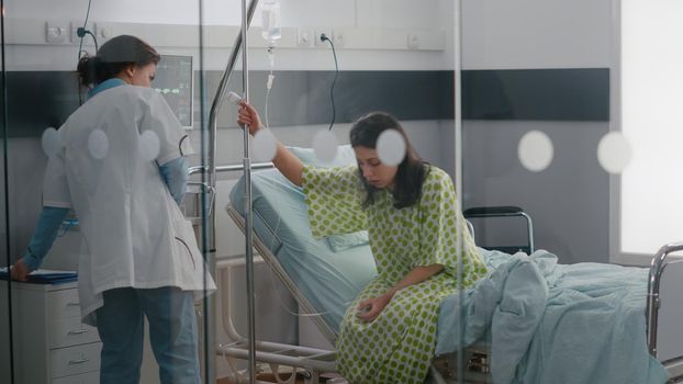 Afro american asisstant with specialist doctor helping patient to stand up from bed holding intravenous IV fluid drip bag after sickness surgery in hospital ward. Practitioner checking cure expertise