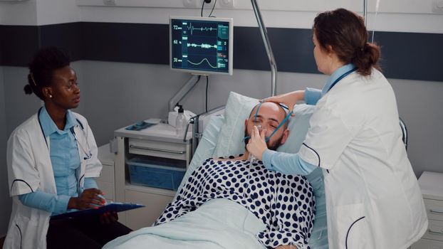 Medical team monitoring heartbeat of sick man during respiratory appointment working in hospital ward. Patient resting in bed while doctor putting oxygen mask checking breath condition