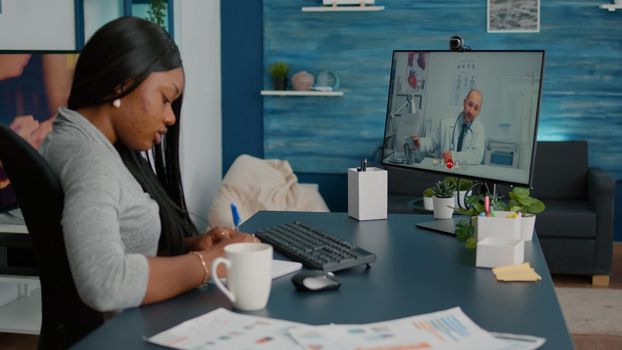 Sick student patient discussing sickness symptoms with physician doctor during online videocall telehealth consultation sitting at desk in living room. Woman writing healthcare treatment on notebook