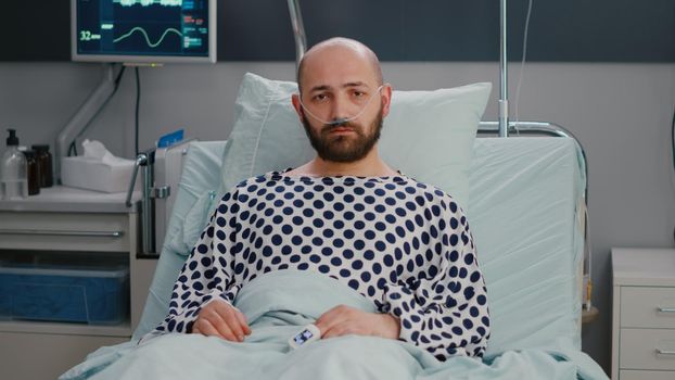 Potrait of hospitalized sick man with nasal oxygen tube having respiratory disorder resting in bed during recovering treatment. Patient waiting for physician while looking at camera in hospital ward