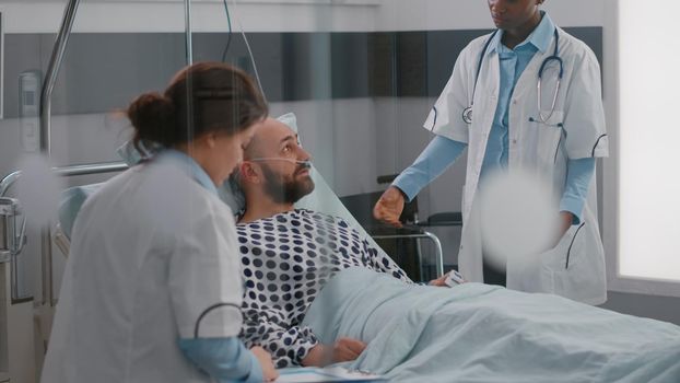 Patient discussing with doctors while lying in bed during sickness recovery in hospital ward. Practitioner writing sickness treatment on clipboard examining medical symptom checking heart pulse