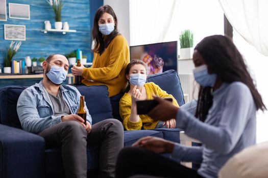 Group of multi ethnic friends hanging out in living room watching clip on smartphone sitting on couch respecting social distancing wearing face mask during global pandemic.