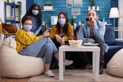 Pov of african woman and friends looking at tv holding remote control wearing face mask because of coronavirus pandemic outbreak drinking beer, relaxing, sitting on sofa in living room.